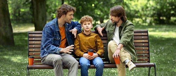Растерянный мальчик сидит на скамейке в парке между двумя взрослыми мужчинами, обсуждающими инвестиции.