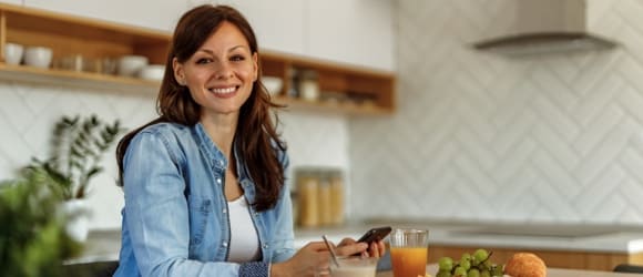 A woman with dark hair is eating breakfast in the kitchen and at the same time is joining III pillar in her phone.