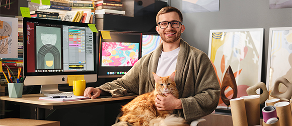 A male designer is sitting at home office, in front of the computer, holding the cat in his arms. 